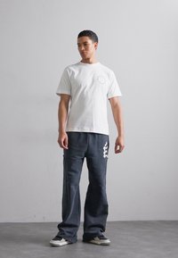 Vit bomull t-shirt med korta ärmar och cirkulär grafik. Mörkgrå vida byxor med vit logotypdetalj. Svart och vit sneakers.