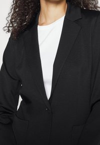 Blazer nero con risvolto, vestibilità su misura e tessuto testurizzato, dotato di due tasche frontali e chiusura a un bottone. Camicia bianca sotto.