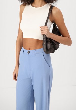Vrouw draagt een wit cropped mouwloos topje, lichtblauwe hoge taille broek en houdt een kleine zwarte schoudertas vast.