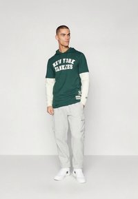 Zelena jopa s kapuco z napisom "New York Yankees" v belem, oblečena čez bež majico z dolgimi rokavi, kombinirana s sivimi hlačami z žepi in belimi supergami.