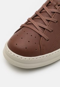 Brown leather sneaker with perforated toe detail, contrasting beige rubber sole, and matching brown laces, featuring a smooth texture.