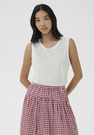 Femme aux longs cheveux noirs portant un haut blanc sans manches et une jupe à carreaux rouges et blancs taille haute, mains dans le dos.