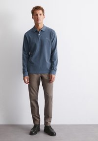 Lange mouw blauwe polo met kraag, gecombineerd met lichtbruine broeken en zwarte schoenen. Het shirt heeft een klein geborduurd logo.