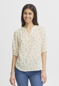 Lichtgele blouse met kleine roze watermeloenpatronen, V-hals, knoopdetail en korte pofmouwen. Zacht, gestructureerd materiaal.