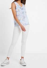 Top senza maniche azzurro chiaro con un motivo floreale, abbinato a jeans skinny bianchi e sneaker bianche. Tessuto liscio, design con scollatura a V.