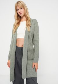 Ανοιχτό πράσινο πλεκτό κασκόλ με μακρύ ανοιχτό μπροστινό μέρος, δύο μπροστινές τσέπες και μακριά μανίκια, φορεμένο πάνω από ένα λευκό crop top και γκρι παντελόνι.