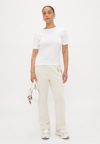 Top corto bianco a maniche corte abbinato a pantaloni sportivi beige e sneakers bianche. Il modello tiene una piccola borsa bianca con un charm decorativo.
