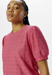 Blouse en dentelle rose avec des manches courtes bouffantes, présentant un motif en diamant complexe et des trous décoratifs à travers le tissu.