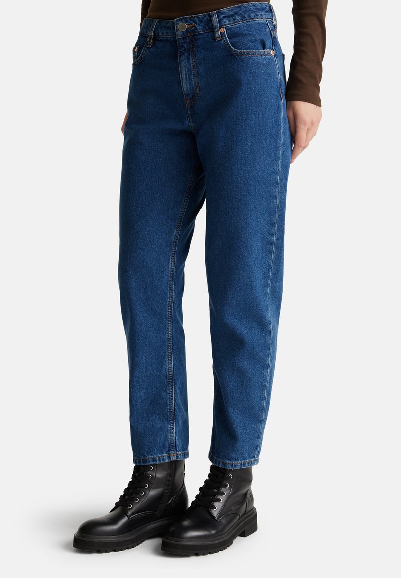 Persona che indossa jeans blu a vita alta e stivaletti neri con lacci, in piedi davanti a uno sfondo chiaro e semplice.