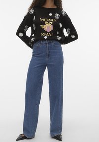 Maglione festivo nero con fiocchi di neve bianchi, testo dorato "MERRY XMAS" e un design di regalo rosa, abbinato a jeans blu a vita alta.