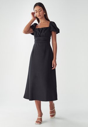 Black midi dress with puff sleeves and a square neckline. Made of smooth fabric, cinched waist, and flowing skirt. Styled with white strappy sandals.