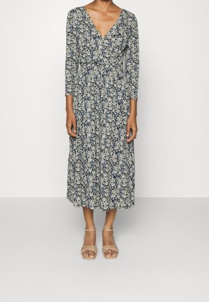 Kobieta w długiej sukience midi z długim rękawem i dekoltem w serek, w niebiesko-białym kwiatowym wzorze, w beżowych sandałkach na obcasie, stojąca na jasnym, jednolitym tle.