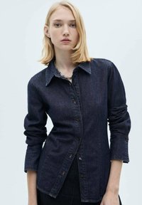 Chemise en denim foncé à boutons, manches longues, col pointu, design ajusté, détails de coutures subtiles, tissu légèrement texturé.