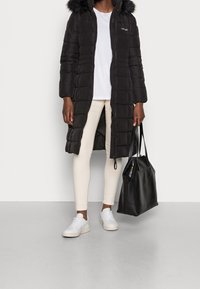 Giacca puffa nera con cappuccio e rifiniture in pelliccia sintetica, abbinata a una t-shirt bianca, leggings crema e sneakers bianche. Inclusa una borsa tote nera.