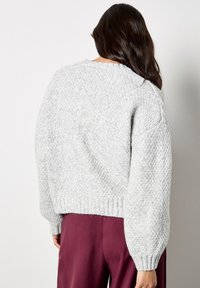 Jasnoszary sweter z dzianiny z oversize'owymi rękawami i ribbowym wykończeniem, z teksturowanym wzorem i luźnym krojem. Noszony z bordowymi spodniami.