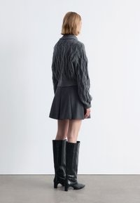 Grau Kabelstrickpullover mit einem cropped Design, kombiniert mit einem dunklen plissierten Rock und knielangen schwarzen strukturierten Lederstiefeln.