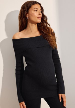 Schwangere Frau mit langen lockigen Haaren, die einen schwarzen, schulterfreien Rippsweater und schwarze Hosen trägt, schaut seitlich vor schlichtem Hintergrund.