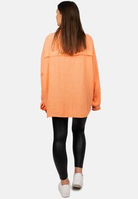 Camicia oversize a maniche lunghe realizzata in tessuto arancione testurizzato, abbinata a leggings neri e scarpe da ginnastica bianche. Presenta un dettaglio sullo spacco posteriore.