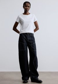 Fehér pamut póló kis lila logóval, bő szabású sötét farmernadrággal párosítva, elöl zsebekkel, szürke varrással és övvel.