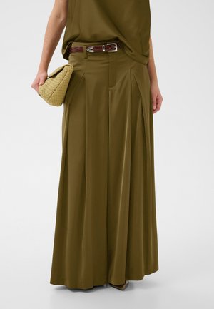 Frau trägt olivgrüne, weit geschnittene Hose und passendes Oberteil, hält eine beige gewebte Clutch mit sichtbarem braunem Gürtel.