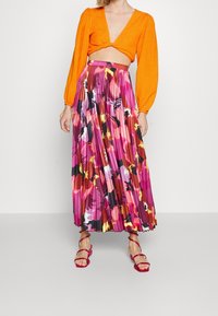 Oranžový žebrovaný crop top s dlouhými rukávy, spojený s plisovanou květovanou maxi sukní v růžové, fialové a žluté barvě. Růžové sandály na tenkých páscích.