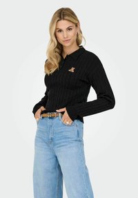 Moteris su ilgais šviesiais plaukais, vilkinti juodą mezginį megztinį su meškos aplikacija ir šviesiai mėlynomis aukšto liemens džinsomis, su smėlio spalvos diržu.