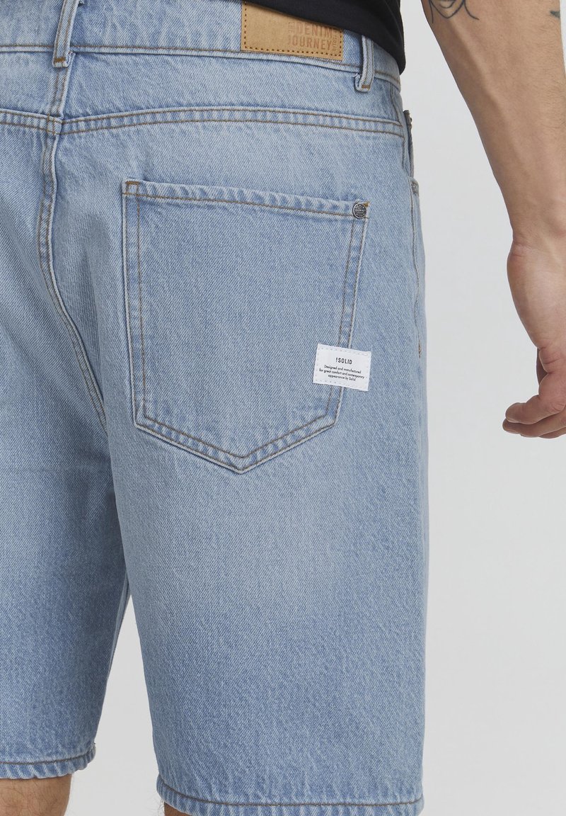 Shorts en jean bleu clair avec une poche arrière et une petite étiquette de marque blanche, portés par une personne avec un bras tatoué visible.