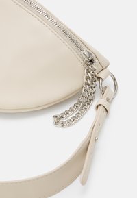Borsa in pelle beige con forma triangolare, dotata di una cerniera argentata, dettaglio in catena metallica e tracolla rimovibile. Texture liscia.