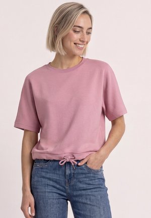 Lachende vrouw met kort blond haar, die een roze top met korte mouwen en een trekkoord in de taille draagt, en een blauwe spijkerbroek, hand in de zak.