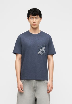 Muž oblečený v tmavomodrom tričku s krátkym rukávom s kvetinovým vzorom na ľavej náprsnej vrecku a svetlosivých nohaviciach.