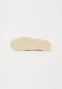 Λευκό αθλητικό παπούτσι με λαστιχένια σόλα, που διαθέτει υφασμένο, σχεδιασμένο πέλμα. Μινιμαλιστικός σχεδιασμός με αντίθετη λεπτομέρεια λογοτύπου στο πλάι.