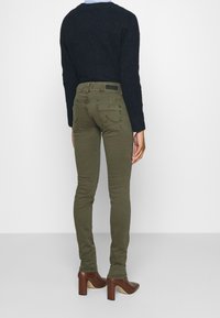 Olivgröna skinny jeans med bakfickor, tillsammans med en svart texturerad tröja, avbildad från baksidan. Bruna klackskor synliga.