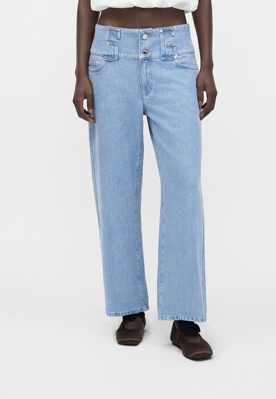 Personne portant un jean taille haute bleu clair à jambes larges avec fermeture double bouton, chaussures marron foncé à bout carré et chaussettes noires.