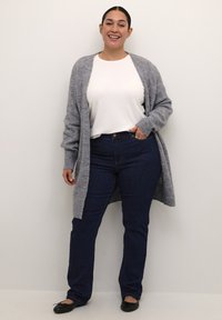 Grau gestrickte cardigan über einem weißen Oberteil, kombiniert mit dunkelblauen Jeans und schwarzen Ballerinas. Das Modell steht lächelnd vor einem neutralen Hintergrund.