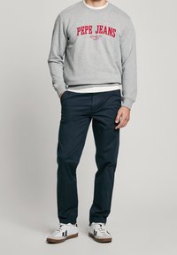 Šedá mikina s červeným logem "PEPE JEANS", nošená přes bílou košili, v kombinaci s tmavě modrými kalhotami a bílými teniskami s hnědými podrážkami.