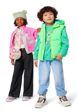 Deux enfants se tiennent côte à côte. L'un porte une veste fuzzy rose, un T-shirt gris avec des graphiques et un jean large noir. L'autre porte un gilet vert fluo, un sweat à capuche assorti et un jogging bleu clair, tous deux en baskets.