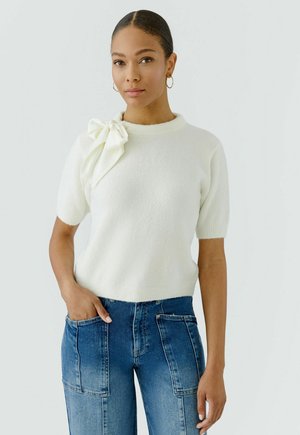 Vrouw die een witte trui met korte mouwen en een strik op de schouder draagt, en blauwe high-waisted jeans, staat met één hand in een zak.
