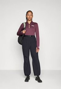 Długi rękaw, top na zamek w ciemnym fioletowym kolorze z fakturowanym wzorem. Luźne ciemne spodnie z kieszeniami i czarny plecak. Czarne buty.