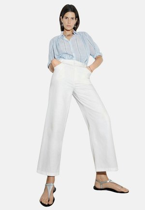 Pantalones anchos blancos con bolsillos frontales, combinados con una blusa ligera de rayas azul claro y mangas cortas. La modelo lleva sandalias plateadas.