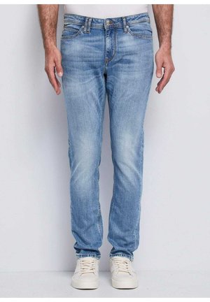 Uomo con jeans blu chiaro e sneakers bianche, in piedi contro uno sfondo grigio chiaro uniforme.