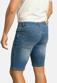 Pantalones cortos de mezclilla en azul medio con un acabado desgastado, con pierna recta, cinco bolsillos y un parche de cuero marrón en la parte trasera de la cinturilla.