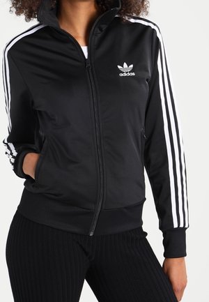 Osoba nosząca czarną kurtkę Adidas z białymi paskami na rękawach i logo, ręce w kieszeniach, w połączeniu z czarnymi prążkowanymi spodniami.