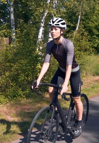Black road bike with slim tires, rider wearing a fitted black and gray cycling jersey and shorts, black helmet, riding on a path surrounded by greenery.