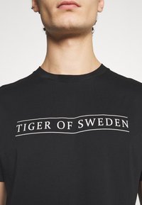 Muž oblečený v černém tričku s bílým nápisem "TIGER OF SWEDEN" napříč hrudníkem, zaměřující se na krk a horní část trupu.