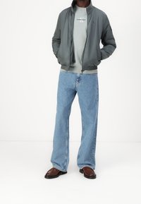 Graue Bomberjacke über einem hellgrauen Pullover, kombiniert mit weit geschnittenen blauen Jeans und braunen Lederschuhen. Einfaches Design mit gerippten Bündchen.