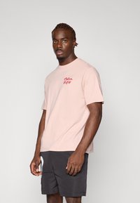 Lyserød bomulds t-shirt med korte ærmer, med røde broderede bogstaver på venstre bryst. Båret med mørke shorts.