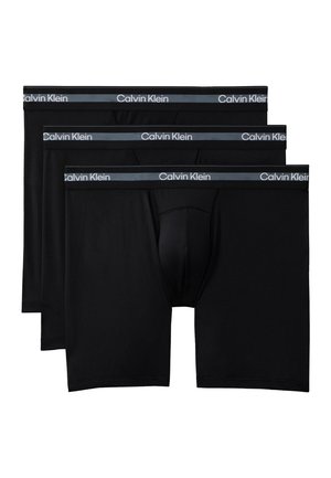 Muskeløs mand stående barfodet mod en ensfarvet baggrund iført sorte Calvin Klein boxershorts med logo i taljebåndet.