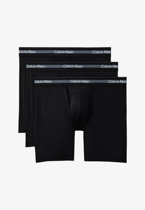 Muskulózny muž stojí bosý na jednoliatom pozadí, oblečený v čiernych boxerkách Calvin Klein s logom na páse.