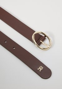 Brauner Ledergürtel mit einer goldenen, kreisförmigen Schnalle mit gedrehtem Design. Das Ende des Gürtels zeigt ein kleines geprägtes Logodetail.