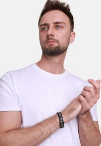 En svart og sølvfarget flettearmbånd på et håndledd. Armbåndet har et kraftig design med en teksturert overflate, anbrakt på en hvit t-skjorte.